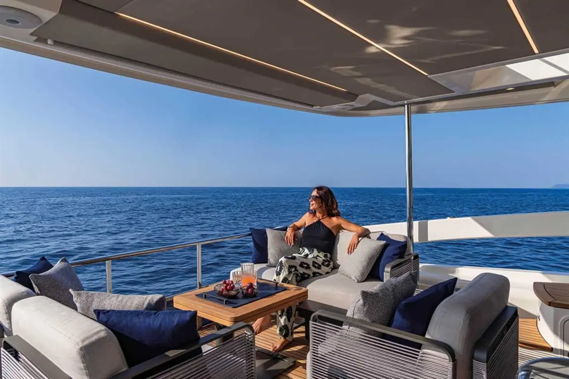 A woman relaxes on a yacht's deck, overlooking the sea, with a wooden table set with drinks and fruit nearby.
